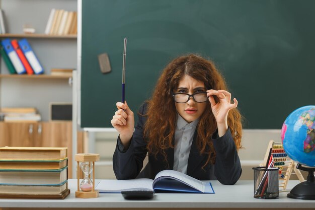 estricta mirando a la cámara joven maestra con gafas sosteniendo un puntero sentado en el escritorio con herramientas escolares en el aula