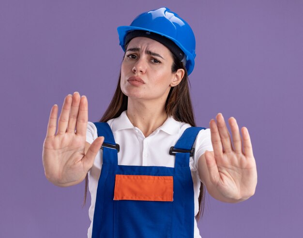 Estricta mujer joven constructor en uniforme mostrando gesto de parada aislado en la pared púrpura