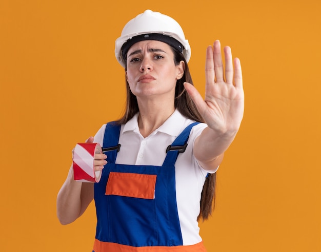 Foto gratuita estricta mujer joven constructora en uniforme sosteniendo cinta adhesiva y mostrando gesto de parada aislado en la pared naranja