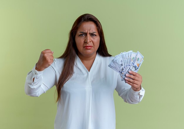 Estricta mujer de mediana edad caucásica casual sosteniendo dinero en efectivo y levantando el puño aislado en la pared verde oliva