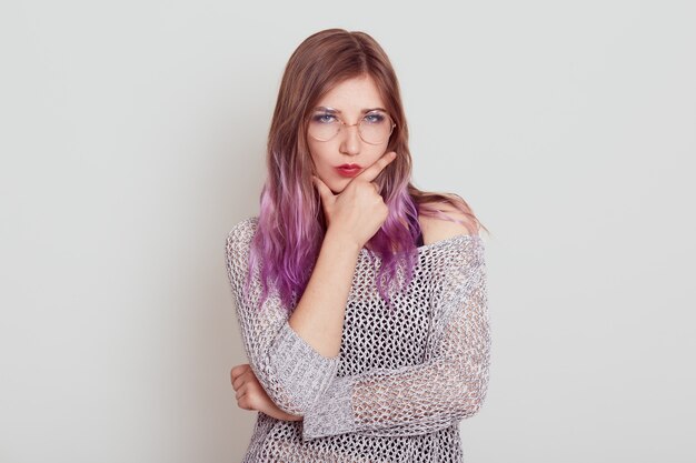 Estricta mujer seria con cabello lila manteniendo los dedos en la barbilla, pensando en cosas importantes o problemas, vistiendo una camisa elegante, posando aislada sobre una pared gris.