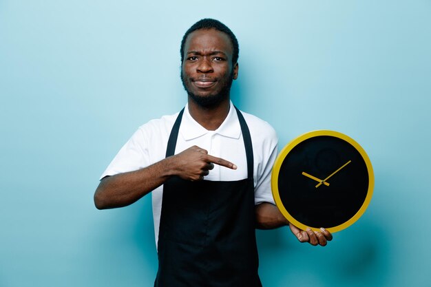 Estricta sujeción y puntos en el reloj de pared joven barbero afroamericano en uniforme aislado sobre fondo azul.