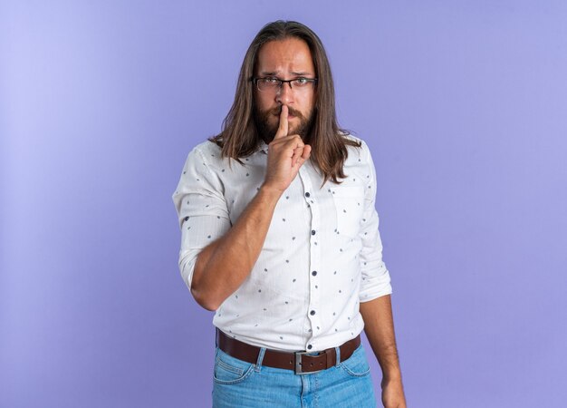 Estricto apuesto hombre adulto con gafas mirando a la cámara haciendo gesto de silencio aislado en la pared púrpura con espacio de copia