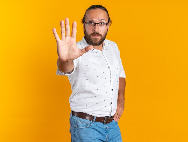 Estricto apuesto hombre adulto con gafas de pie en la vista de perfil manteniendo la mano en la cintura haciendo gesto de parada