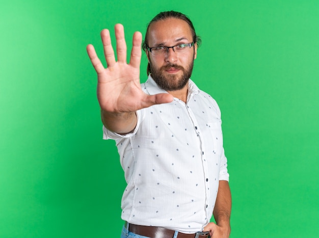 Estricto apuesto hombre adulto con gafas de pie en la vista de perfil manteniendo el pulgar en el cinturón haciendo gesto de parada