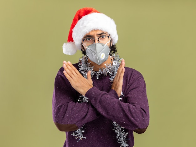 Estricto chico guapo joven con sombrero de navidad y máscara médica con guirnalda en el cuello mostrando gesto de no aislado sobre fondo verde oliva