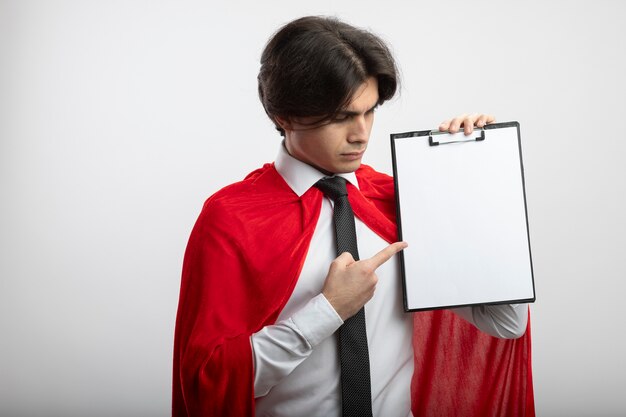 Estricto chico joven superhéroe con corbata sosteniendo y apunta al portapapeles aislado sobre fondo blanco.