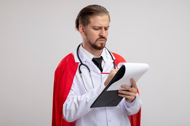 Estricto chico joven superhéroe vistiendo bata médica con estetoscopio escribiendo algo en el portapapeles aislado en blanco