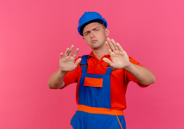 Estricto constructor masculino joven con uniforme y casco de seguridad mostrando gesto de parada en rosa