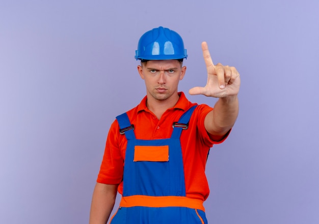 Estricto constructor masculino joven con uniforme y casco de seguridad mostrando gesto en púrpura