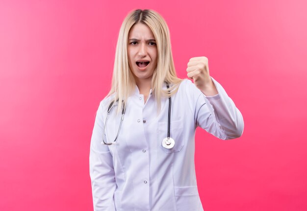 Estricto doctor joven rubia con estetoscopio en bata médica puño levantado sobre fondo rosa aislado