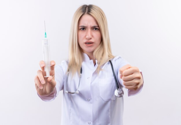 Foto gratuita estricto doctor joven rubia con estetoscopio y bata médica sosteniendo la jeringa sosteniendo el puño a la cámara sobre fondo blanco aislado