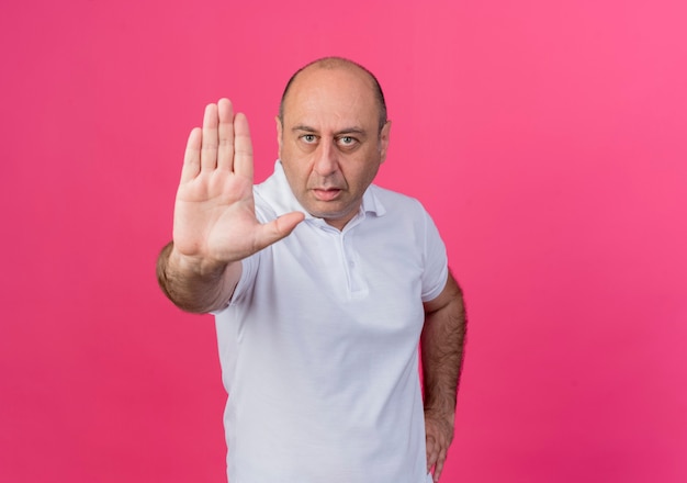 Estricto empresario maduro casual manteniendo la mano en la cintura y haciendo gesto de parada en la cámara aislada sobre fondo rosa con espacio de copia