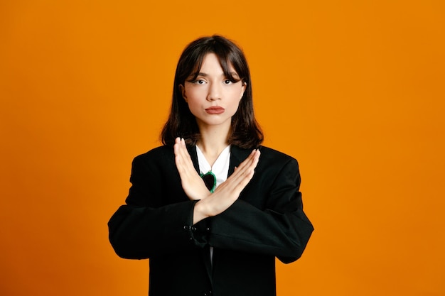Estricto gesto mostrando joven hermosa mujer vistiendo chaqueta negra aislado sobre fondo naranja