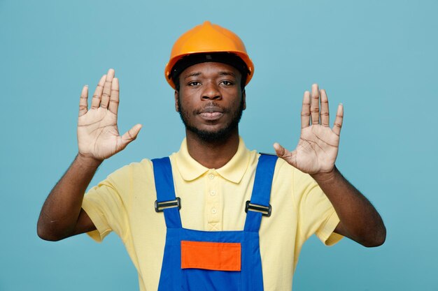 Estricto gesto de parada joven constructor afroamericano en uniforme aislado sobre fondo azul.