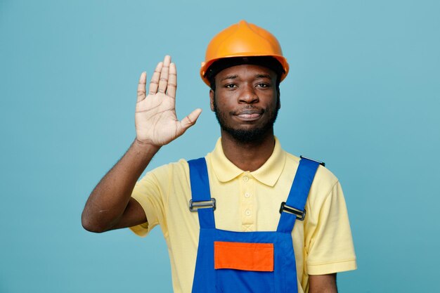 Estricto gesto de parada joven constructor afroamericano en uniforme aislado sobre fondo azul.