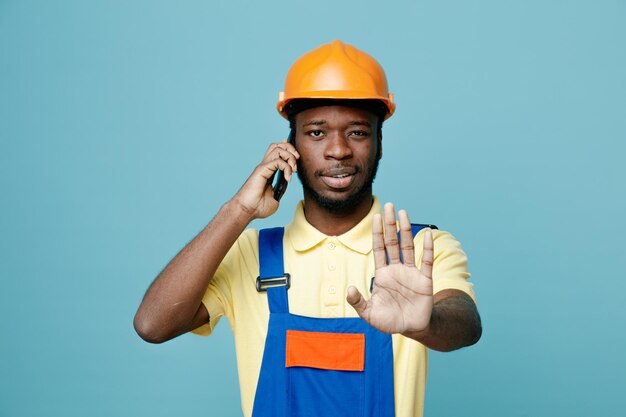 Estricto gesto de parada joven constructor afroamericano en uniforme habla por teléfono aislado sobre fondo azul.