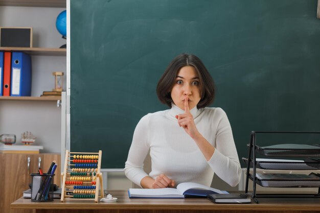 estricto gesto de silencio joven maestra sentada en el escritorio con herramientas escolares en el aula