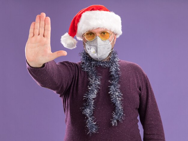 Estricto hombre de mediana edad con gorro de Papá Noel y máscara protectora con guirnalda de oropel alrededor del cuello con gafas haciendo gesto de parada aislado en la pared púrpura