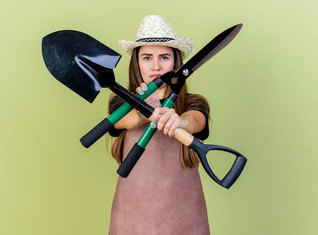 Foto gratuita estricto jardinero hermosa chica en uniforme con sombrero de jardinería sosteniendo y cruzando las tijeras con pala aislado sobre fondo verde oliva