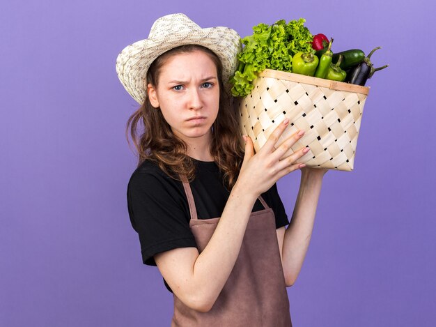 Estricto jardinero joven vistiendo sombrero de jardinería sosteniendo canasta de verduras en el hombro