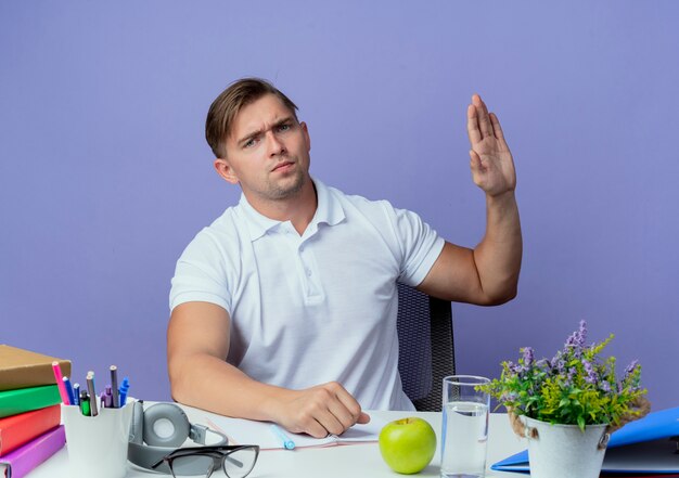 Estricto joven apuesto estudiante sentado en el escritorio con herramientas escolares extendiendo la mano al lado aislado en azul
