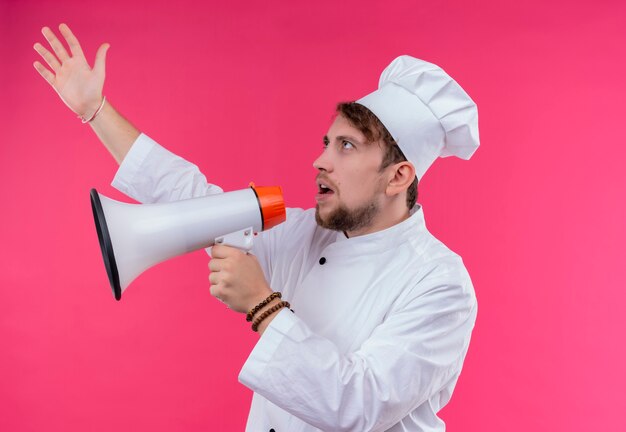 Un estricto joven chef barbudo con uniforme blanco hablando por megáfono con la mano levantada sobre una pared rosa