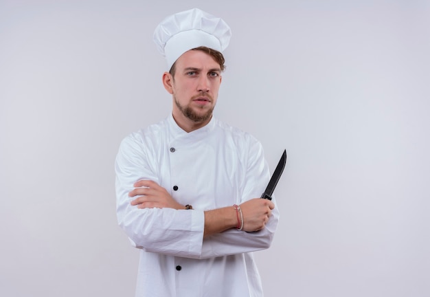 Un estricto joven chef barbudo con uniforme blanco sosteniendo un cuchillo con las manos juntas mientras mira en una pared blanca