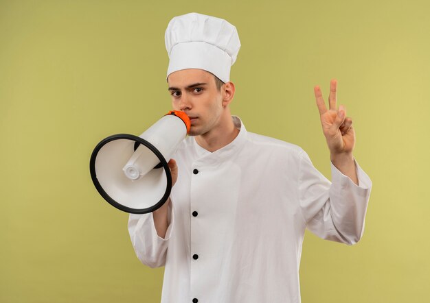Estricto joven cocinero vistiendo uniforme de chef habla en altavoz mostrando gesto de paz