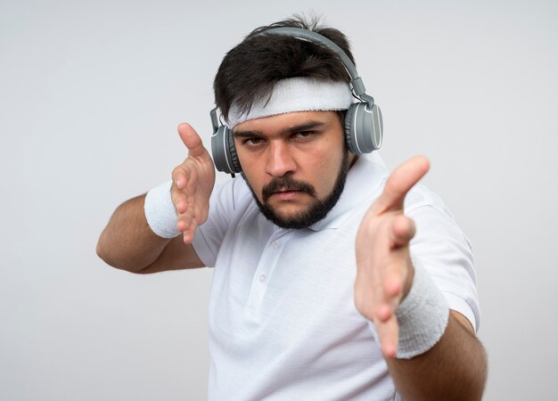 Estricto joven deportivo con diadema y muñequera con auriculares sosteniendo las manos aisladas en la pared blanca