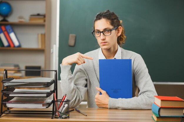 Estricto joven maestro sosteniendo y apuntando al cuaderno sentado en el escritorio con herramientas escolares en el aula