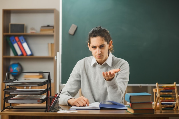 Foto gratuita estricto joven maestro sosteniendo la mano frente a la cámara sentado en el escritorio con herramientas escolares en el aula