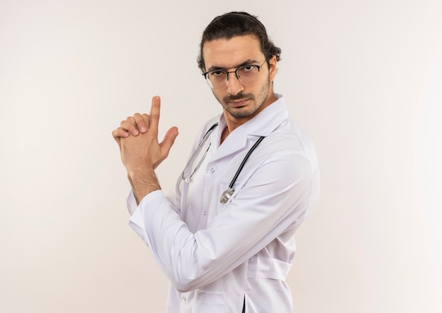 Estricto joven médico con gafas ópticas vistiendo túnica blanca con estetoscopio sgowing gesto de pistola en pared blanca aislada con espacio de copia