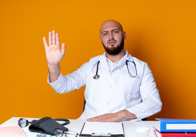 Estricto joven médico varón calvo vistiendo bata médica y estetoscopio sentado en el escritorio con herramientas médicas que muestran gesto de parada aislado en la pared naranja