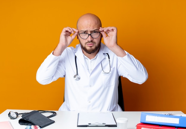 Foto gratuita estricto joven médico varón calvo vistiendo bata médica y un estetoscopio sentado en el escritorio con herramientas médicas toman gafas en naranja