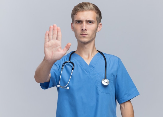 Estricto joven médico vistiendo uniforme médico con estetoscopio mostrando gesto de parada aislado en la pared blanca