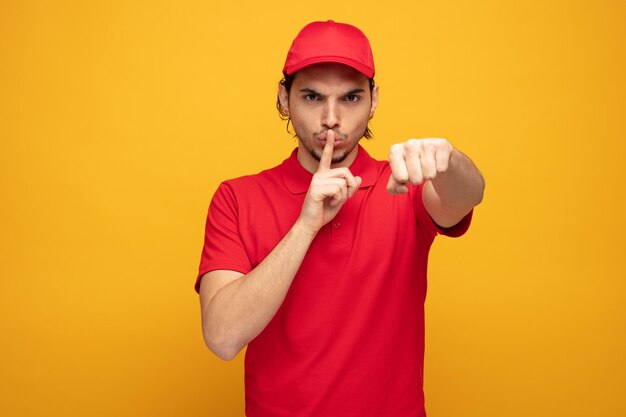 estricto joven repartidor con uniforme y gorra mirando a la cámara mostrando un gesto de silencio estirando el puño hacia la cámara aislada en el fondo amarillo