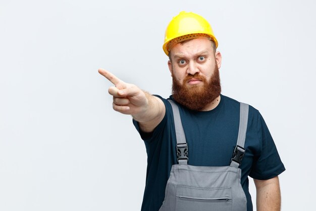 Estricto joven trabajador de la construcción con casco de seguridad y uniforme mirando a la cámara apuntando al lado aislado en fondo blanco con espacio para copiar