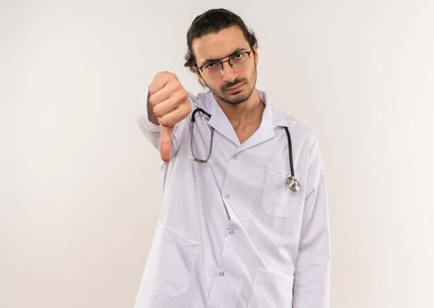 Estricto médico varón joven con gafas ópticas vistiendo túnica blanca con estetoscopio con el pulgar hacia abajo