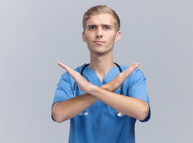 Estricto médico varón joven vistiendo uniforme médico con estetoscopio mostrando gesto de no aislado en la pared blanca