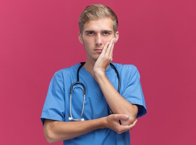 Estricto médico varón joven vistiendo uniforme médico con estetoscopio poniendo la mano en la mejilla aislada en la pared rosa con espacio de copia
