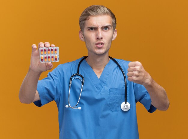 Estricto médico varón joven vistiendo uniforme médico con estetoscopio sosteniendo pastillas aisladas en la pared naranja