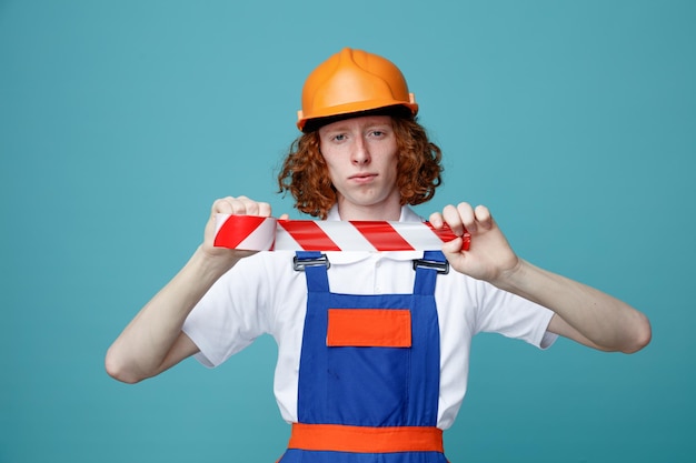 Estricto mirando a la cámara joven constructor hombre en uniforme sosteniendo cinta adhesiva aislado sobre fondo azul.