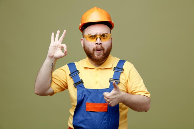 Estricto mostrando diferentes gestos joven constructor hombre en uniforme aislado sobre fondo verde