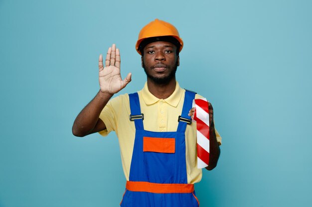 Estricto mostrando gesto de parada sosteniendo cinta adhesiva joven constructor afroamericano en uniforme aislado sobre fondo azul.