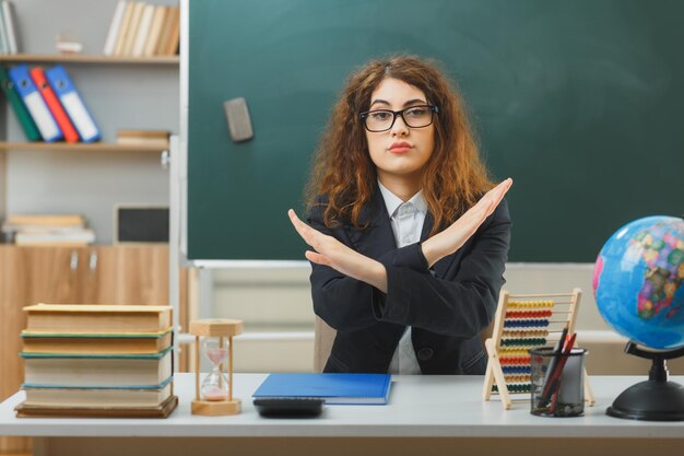 estricto sin mostrar ningún gesto joven maestra con gafas sentada en el escritorio con herramientas escolares en el aula