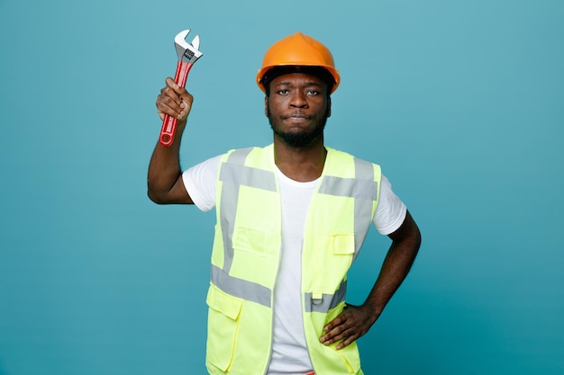 Estricto poner la mano en las caderas joven constructor afroamericano en uniforme con llave de gas aislado sobre fondo azul.