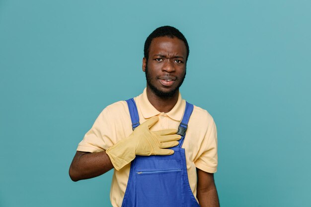 Estricto poner la mano en el corazón joven limpiador afroamericano macho en uniforme con guantes aislado sobre fondo azul.