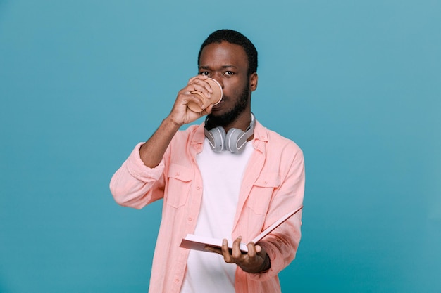 Estricto sujetando una taza de café con un libro joven afroamericano usando audífonos en el cuello aislado de fondo azul