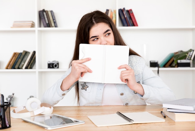 Estudiante adolescente con libro abierto en la cara sentado en la mesa